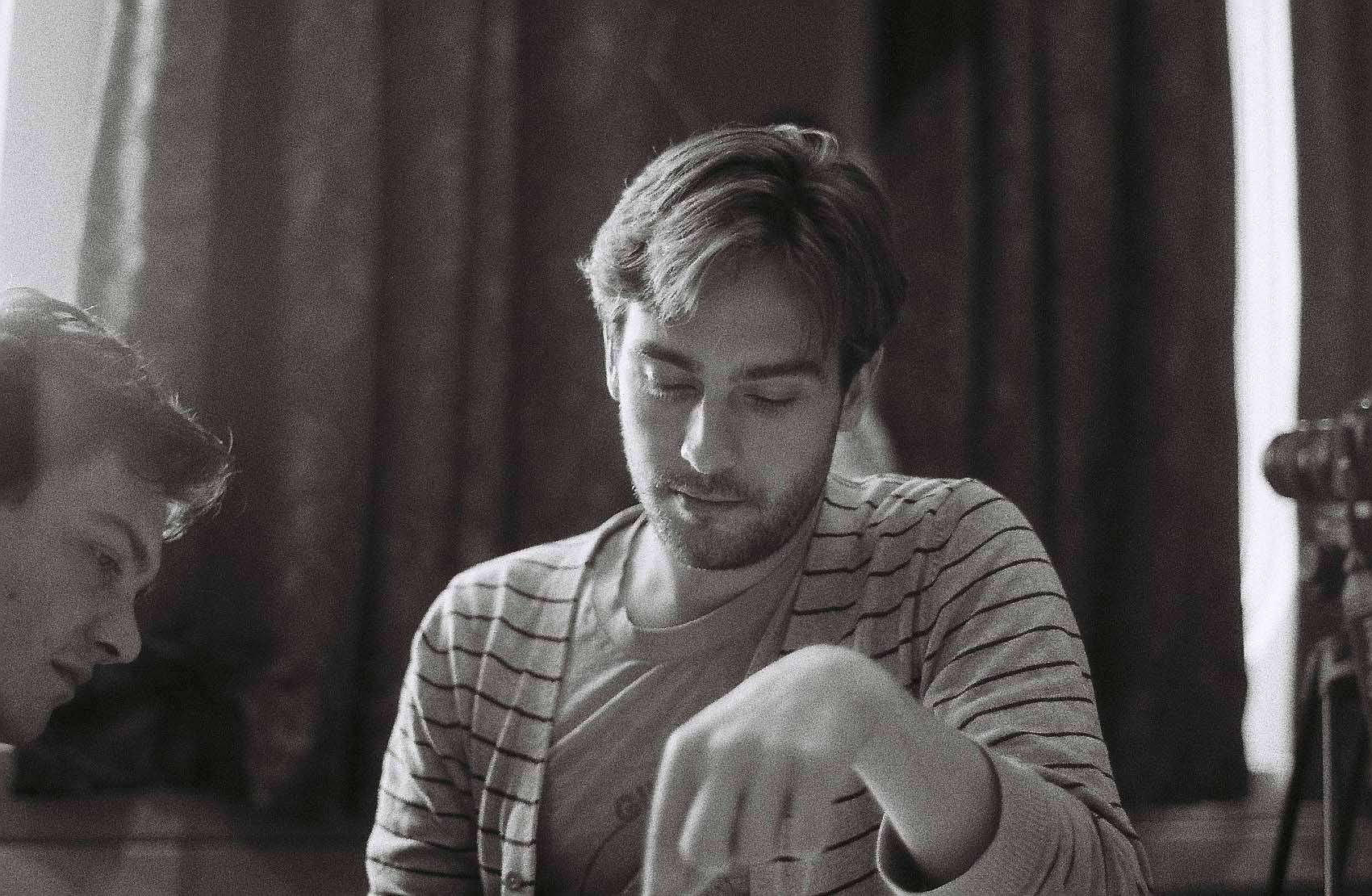 Black and white photo of Tom Mison in striped cardigan, looking down 