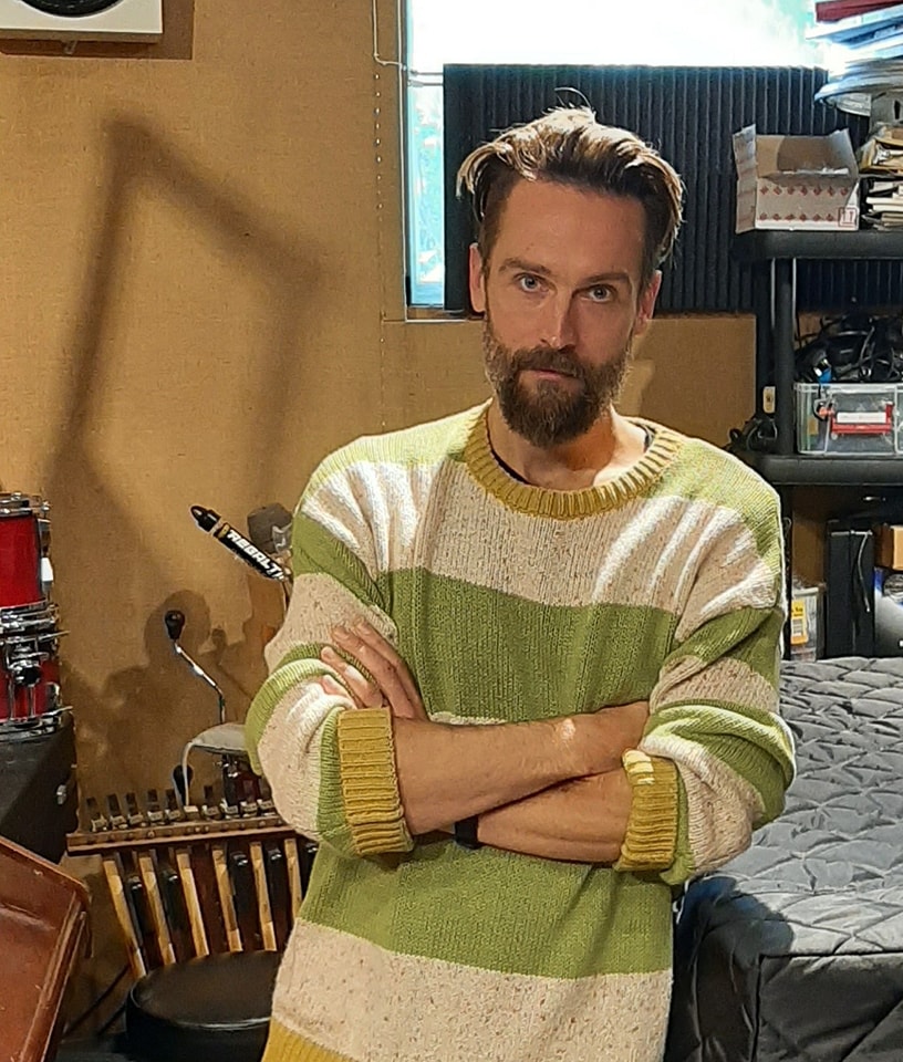 Tom stands with arms crossed, wearing a sweater